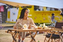Schöne ethnische Asiatin mit Sonnenbrille sitzt am Tisch, während sie im Urlaub eine entspannte Zeit auf dem Campingplatz verbringt und wegschaut — Stockfoto
