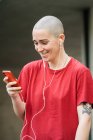Giovane donna omosessuale felice in t-shirt e auricolari con cellulare guardando lo schermo mentre ascolta musica — Foto stock