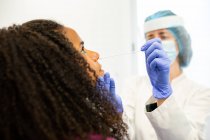 Женщина-врач в защитной форме, латексных перчатках и маске для лица делает тест на носовую маску с ватным бутоном у афроамериканской пациентки в клинике во время вспышки коронавируса — стоковое фото