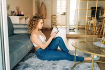 Ganzkörper-Seitenansicht einer positiven jungen Frau mit lockigem Haar in lässiger Kleidung und Hausschuhen, die auf einem Teppich neben dem Sofa sitzt und am Wochenende zu Hause interessante Bücher liest — Stockfoto