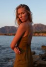 Vue latérale de la jeune femme debout sur la côte contre la mer bleue agitant au coucher du soleil et regardant la caméra — Photo de stock