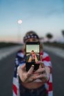 Патріотична американська жінка, загорнута в національний прапор США, робить сельфі на мобільному телефоні, стоячи на дорозі — стокове фото