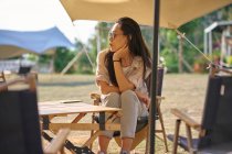Hermosa mujer asiática étnica en gafas de sol sentada en la mesa mientras disfruta de un momento de relax en la zona de acampada durante las vacaciones mirando hacia otro lado - foto de stock