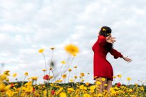 Снизу вид на модную женщину в красном сарафане и с цветочной короной, стоящей с закрытыми глазами на цветущем поле с желтыми и красными цветами с протянутыми руками в теплый летний день — стоковое фото