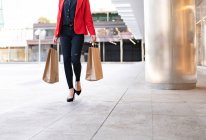 Crop comprador femenino con bolsas de papel y café para llevar de pie cerca de escaparate de la tienda en la ciudad - foto de stock