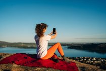 Обратный вид на неузнаваемую женщину-туристку с вьющимися волосами в повседневной одежде, делающую селфи, сидящее на одеяле на скалистом утесе над морем в солнечный день — стоковое фото