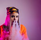 Cool femelle en tenue de rue fumer e cigarette et expirer la fumée par le nez sur fond violet en studio avec éclairage au néon rose — Photo de stock