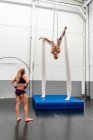 Full body fit female in sportswear looking at muscular male partner performing upside down exercises on aerial silks while training together in modern fitness center — Stock Photo