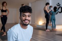 Sorridente maschio afroamericano in activewear seduto sul tappeto e guardando la fotocamera dopo lezione di yoga sullo sfondo di diverse persone — Foto stock