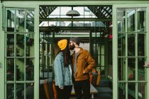 Alegre pareja de hipsters de moda de pie en una terraza de madera mientras se toman de la mano y miran por encima del hombro - foto de stock