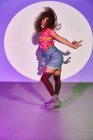 Вид сбоку афроамериканской танцовщицы в шортах, танцующей, глядя в камеру с потухшим языком, стоя в неоновом свете в студии — стоковое фото