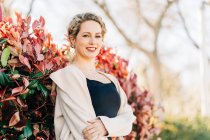 Glückliche blonde Frau in elegantem Kleid und Mantel, die zwischen Bäumen steht und in die Kamera blickt — Stockfoto