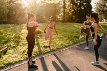 Young multiracial female athletes in sportswear stretching arm and legs on asphalt footpath in town on sunny day — Stock Photo