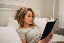 Young female with curly hair lying on bed reading book during weekend at home — Stock Photo