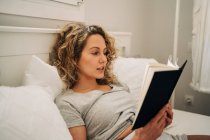 Young female with curly hair lying on bed reading book during weekend at home — Foto stock