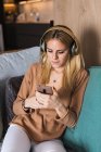 Young female sitting on sofa and enjoying music in headphones while looking at screen of smartphone — Stock Photo
