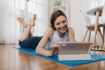 Deliziosa femmina magra sdraiata su tappetino e tablet di navigazione durante la scelta della lezione online per praticare yoga — Foto stock