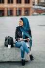 Mulher muçulmana cuidadosa em hijab escrevendo em diário enquanto sentada na rua da cidade e olhando para longe — Fotografia de Stock