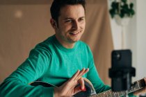 Crop cheerful adult male vlogger with guitar and plectrum recording video tutorial on cellphone at home — Stock Photo