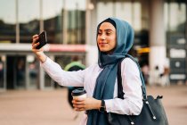 Вид сбоку этнической женщины в хиджабе, стоящей рядом с железнодорожным вокзалом и делающей селфи на мобильном телефоне в ожидании поезда — стоковое фото