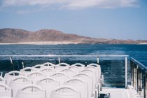 Sillas blancas vacías en la cubierta del barco de crucero navegando en agua azul del mar con la montaña en la orilla - foto de stock