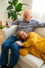 From above of mature female lying on knees of man sitting on sofa in living room and browsing smartphone — Stock Photo