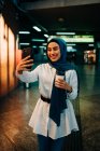 Donna etnica in hijab in piedi sul binario sulla stazione ferroviaria e scattare selfie sul telefono cellulare in attesa di treno — Foto stock