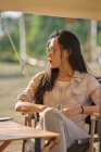 Mulher asiática étnica bonita em óculos de sol sentados à mesa enquanto tendo um tempo relaxante na área de acampamento durante as férias olhando para longe — Fotografia de Stock