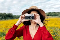 Anonymous smiling female in hat taking photo on vintage camera on meadow under cloudy sky — Stock Photo