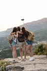 Von hinten reisende Freundinnen mit Rucksäcken, die auf einem Hügel stehen und im Sommer vor dem Hintergrund der Gebirgsketten Selbstaufnahmen mit dem Smartphone machen — Stockfoto
