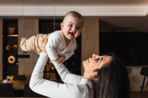 Vista lateral da mãe encantada jogando adorável bebê sorridente enquanto se divertem juntos em casa — Fotografia de Stock