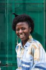 Young African American female with traditional comb on afro hair dressed in stylish colorful informal outfit smiling happily while standing against glass wall with reflection of urban street — Stock Photo