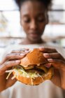 Урожай размыл афроамериканку в повседневной одежде, поедая вкусный свежий чизбургер в светлой комнате. — стоковое фото