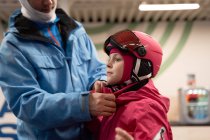 Positive young father in warm activewear and ski balaclava putting protective helmet on cute daughter head in modern ski center — Stock Photo