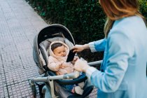 De cima bonito bebê engraçado com smartphone vestindo roupas quentes sentado no carrinho e olhando para a mãe turva na rua primavera — Fotografia de Stock