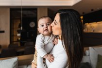 Vista laterale della madre felice che tiene e bacia adorabile bambino sorridente mentre si diverte insieme a casa — Foto stock