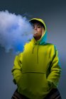Знизу впевнена жіноча хіпстерка в светрі курить електронну сигарету в студії на сірому фоні і дивиться на камеру — стокове фото