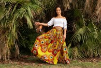 Joven mujer étnica alegre en falda africana ornamental mirando a la cámara contra las plantas de palma en el prado - foto de stock