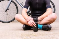 Ground level of crop unrecognizable male cyclist in sportswear with bottle sitting with crossed legs against bike on walkway - foto de stock