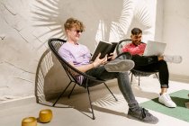 Cuerpo completo de elegantes amigos masculinos multirraciales jóvenes que utilizan el ordenador portátil y libro de lectura mientras se relaja en las sillas en la terraza en el día soleado - foto de stock