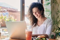 Vista laterale di felice dipendente etnica femminile che lavora su netbook a tavola con delizioso panino vegetariano bagel e bevande — Foto stock