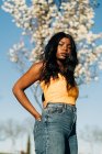 Niedriger Winkel der schönen Afroamerikanerin, die im blühenden Frühlingspark steht und sonniges Wetter genießt, während sie in die Kamera blickt — Stockfoto
