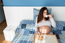 Сверху мягкая беременная женщина сидит на кровати с подносом и пьет кофе с печеньем во время завтрака утром — стоковое фото