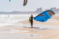 Sportlerin im Neoprenanzug mit aufblasbarem Drachen spaziert am Sandstrand und blickt gegen stürmischen Ozean — Stockfoto