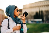 Side view of ethnic female in headscarf and stylish sunglasses standing with takeaway drink on street and recording voice message on mobile phone — Fotografia de Stock