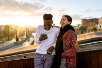 Счастливая многорасовая пара в стильной одежде с мобильным телефоном, говорящая в городе под облачным небом в сумерках — стоковое фото
