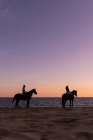Vista laterale di silhouette anonime coppia su giumente contemplando oceano infinito dalla riva sabbiosa al tramonto — Foto stock