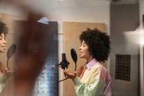 Бічний вид на молоду афроамериканку співачку, що співає мікрофоном, з поп-фільтром у студії звукозапису. — стокове фото