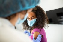 Unrecognizable female medical specialist in protective uniform, latex gloves and face mask vaccinating African American female patient in clinic during coronavirus outbreak — Stock Photo