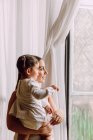 Side view of ethnic mother with cute little daughter looking out of window while standing in room at home — Stock Photo